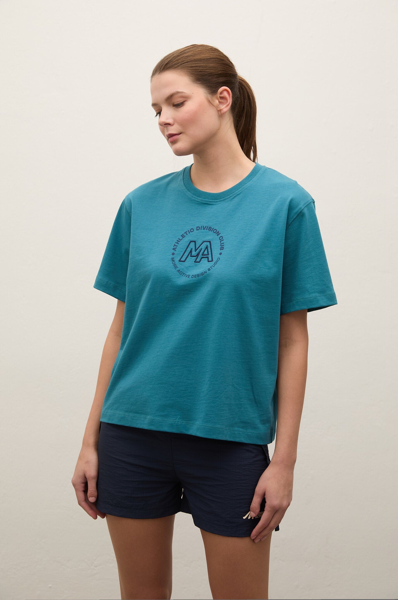 A woman wearing a teal short-sleeve t-shirt with a circular 'AA' logo on the chest, paired with dark navy shorts, standing against a plain off-white wall.
