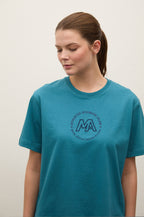 A woman wearing a teal short-sleeve crewneck t-shirt with a dark gray circular graphic on the chest featuring the letters 'MA' and surrounding text, set against a plain off-white background.