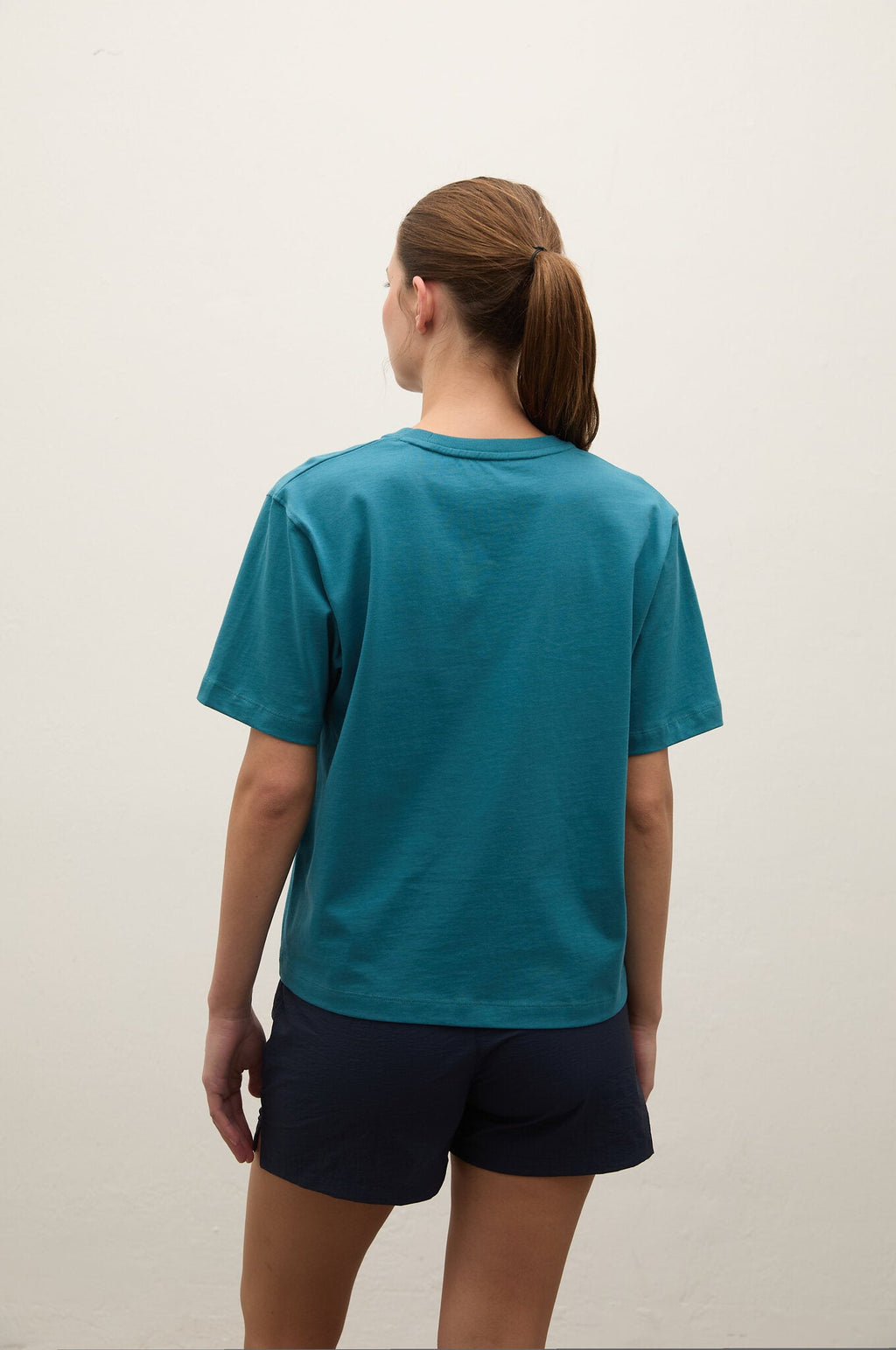 A woman viewed from behind wearing a teal short-sleeve crewneck t-shirt and dark navy shorts, standing against a plain off-white wall.