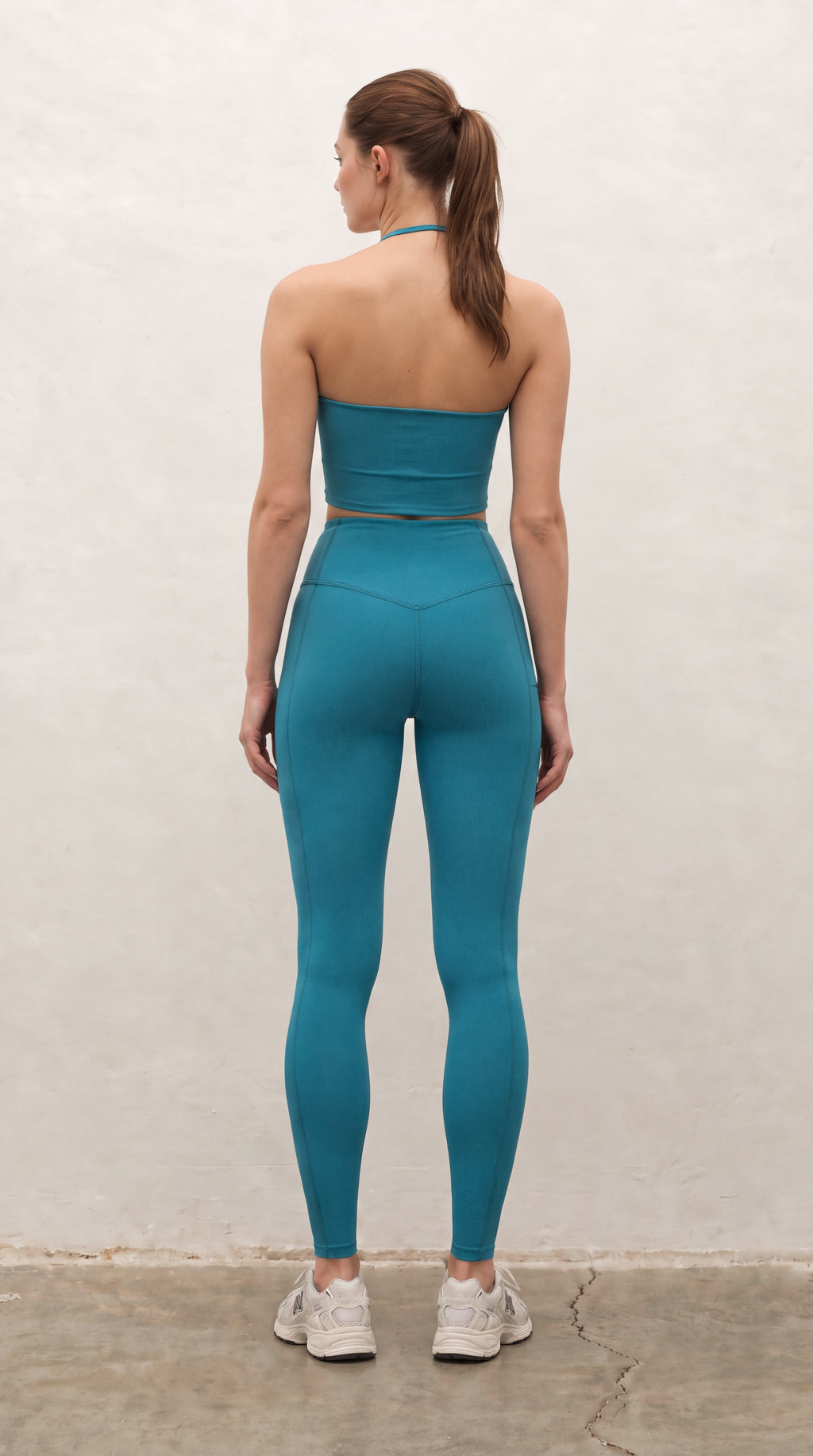 A woman stands with her back to the camera wearing a matching teal blue-green activewear set consisting of a strapless bandeau-style crop top and high-waisted leggings, paired with white sneakers, against a textured off-white wall.