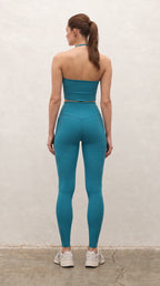 A woman stands with her back to the camera wearing a matching teal blue-green activewear set consisting of a strapless bandeau-style crop top and high-waisted leggings, paired with white sneakers, against a textured off-white wall.
