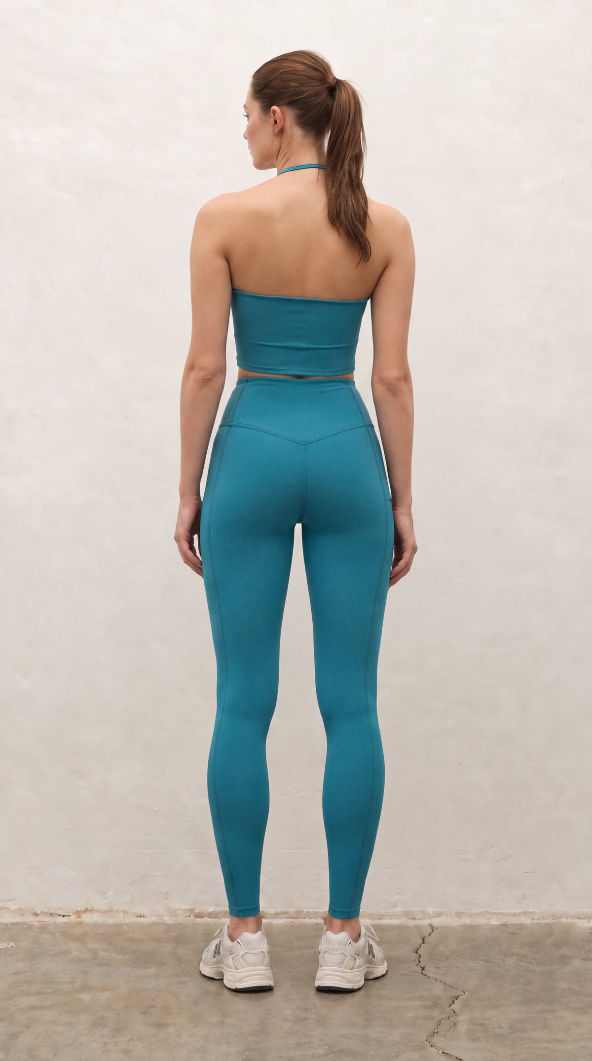 A woman stands with her back to the camera wearing a matching teal blue-green activewear set consisting of a strapless bandeau-style crop top and high-waisted leggings, paired with white sneakers, against a textured off-white wall.