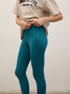 A model wearing high-waisted teal blue leggings and a beige graphic t-shirt, standing against a plain off-white wall.