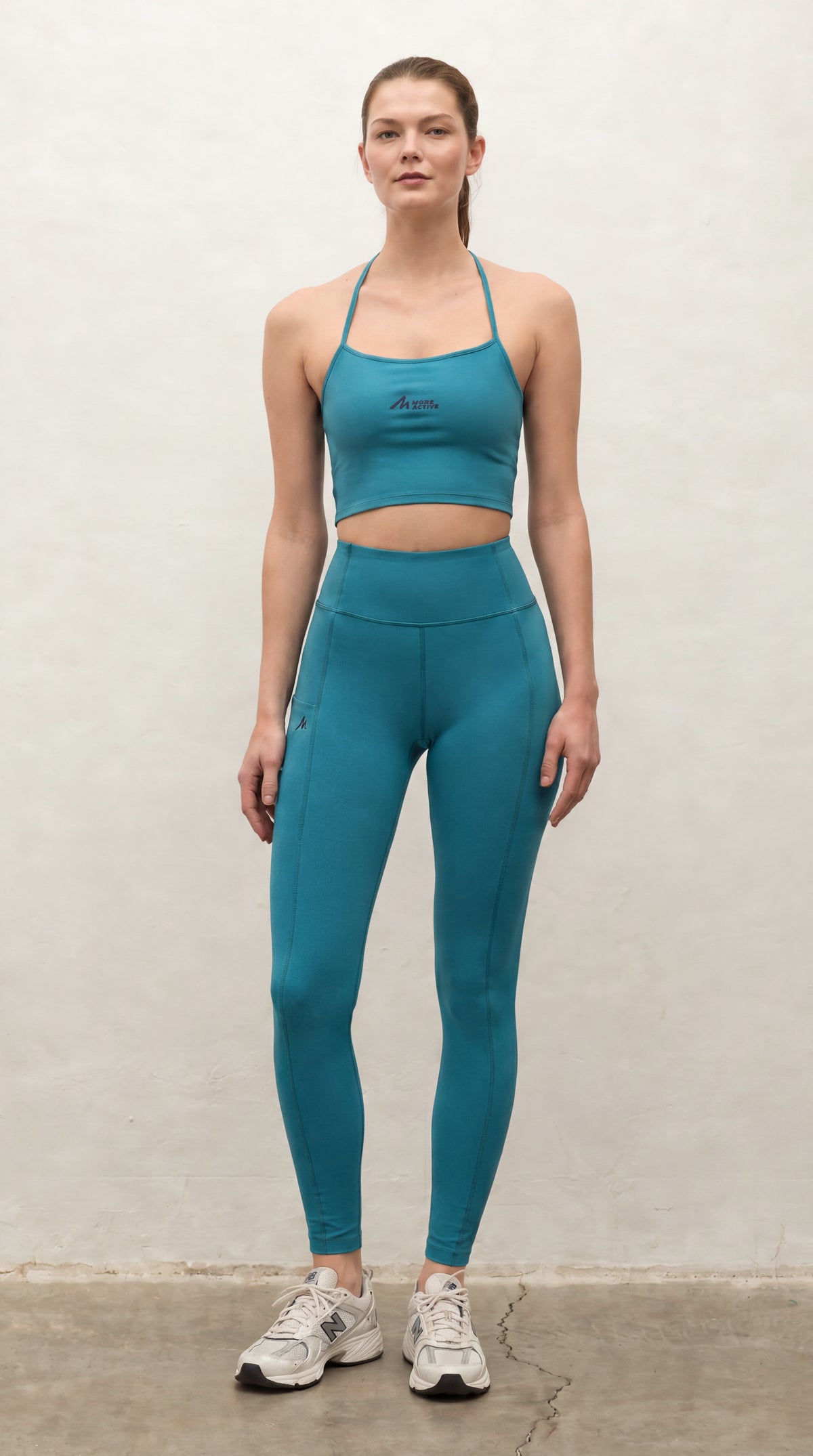 A woman modeling a matching teal blue activewear set consisting of a sports bra and high-waisted leggings, paired with white sneakers, standing against a textured off-white wall.