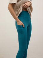 A model wearing high-waisted teal leggings with a side pocket and a small embroidered logo, paired with a beige cropped t-shirt, shown from the waist down against a plain off-white wall.