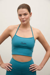 A woman modeling a teal athletic crop top and matching leggings against a plain light background. The crop top features a small black logo on the center chest.