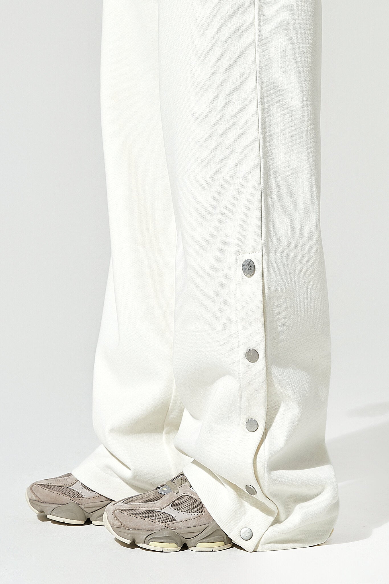 Close-up of white wide-leg trousers with a vertical row of silver buttons down the outer side, shown on a person wearing taupe sneakers against a light gray studio background.