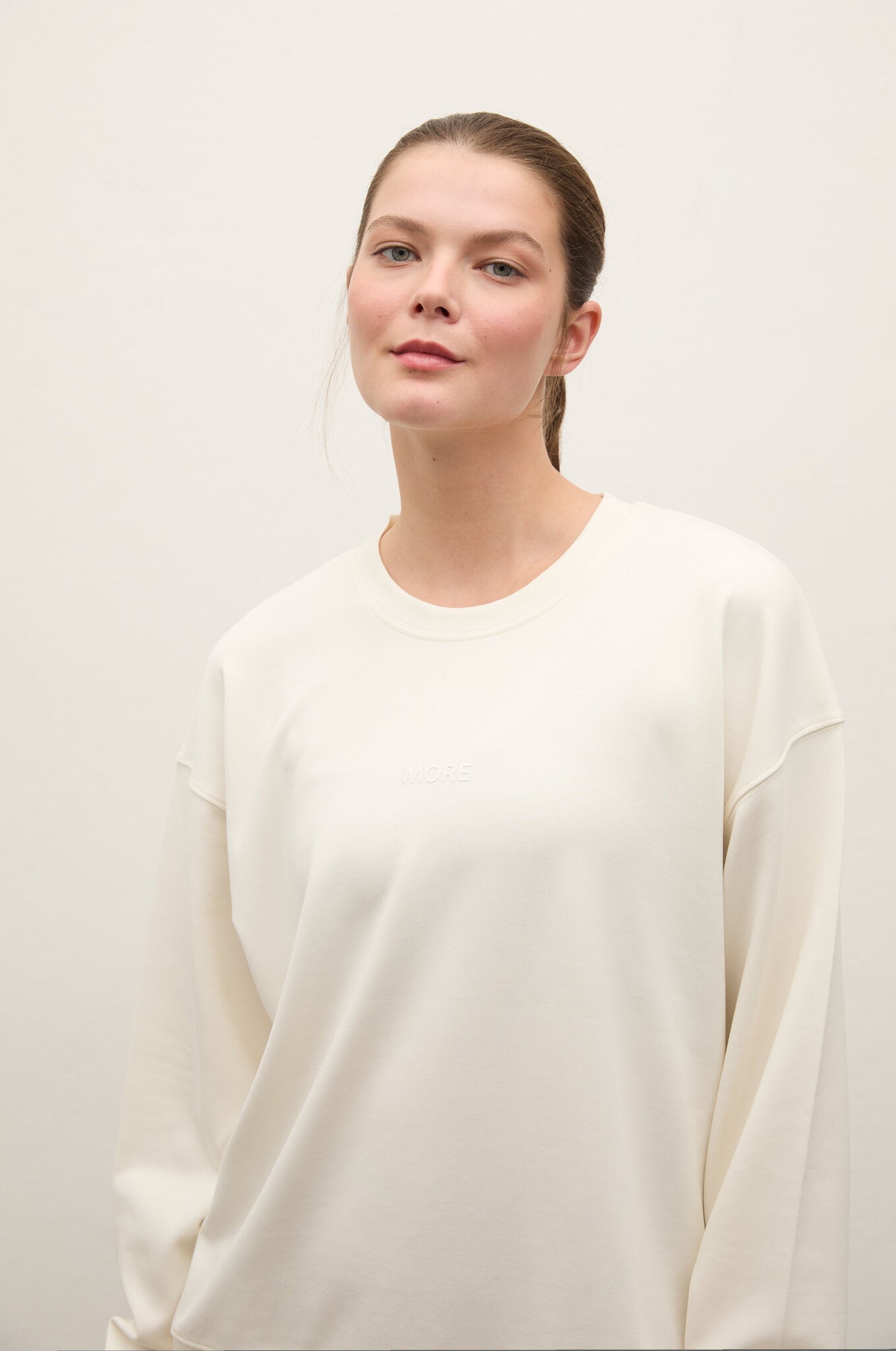 A woman with her hair pulled back wears a cream-colored crewneck sweatshirt against a plain, light off-white background.