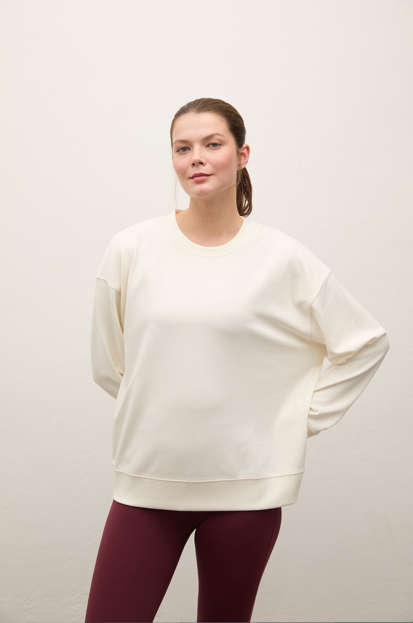 A woman modeling an oversized cream-colored crewneck sweatshirt paired with burgundy leggings, standing against a plain off-white studio background.