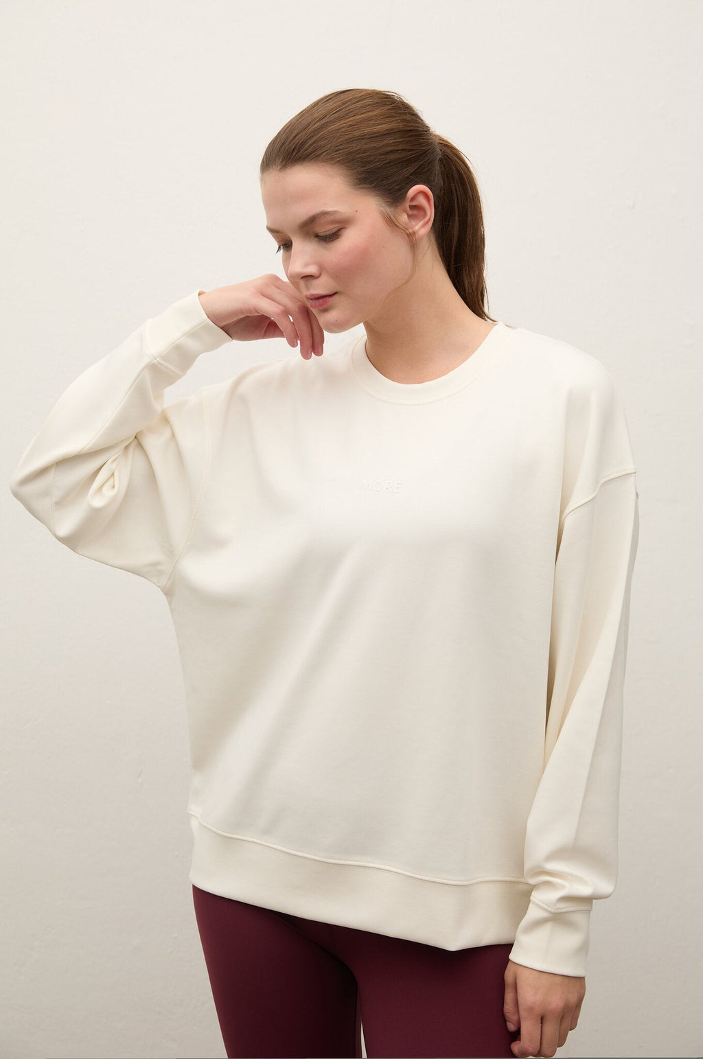 A woman wearing an oversized cream-colored crewneck sweatshirt and burgundy leggings, posing against a plain off-white studio background.