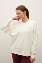 A woman wearing an oversized cream-colored crewneck sweatshirt and burgundy leggings, posing against a plain off-white studio background.