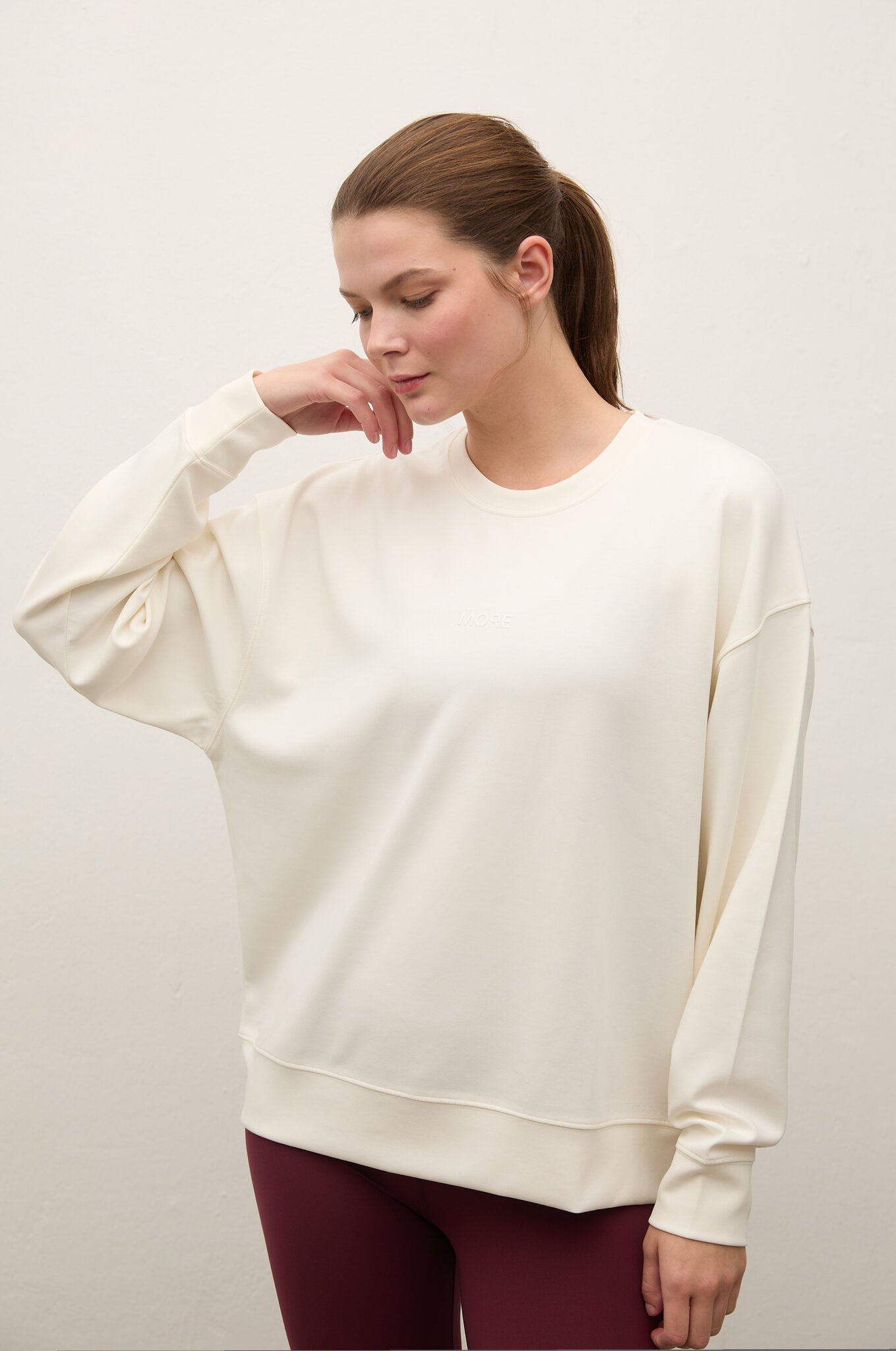 A woman wearing an oversized cream-colored crewneck sweatshirt and burgundy leggings, posing against a plain off-white studio background.