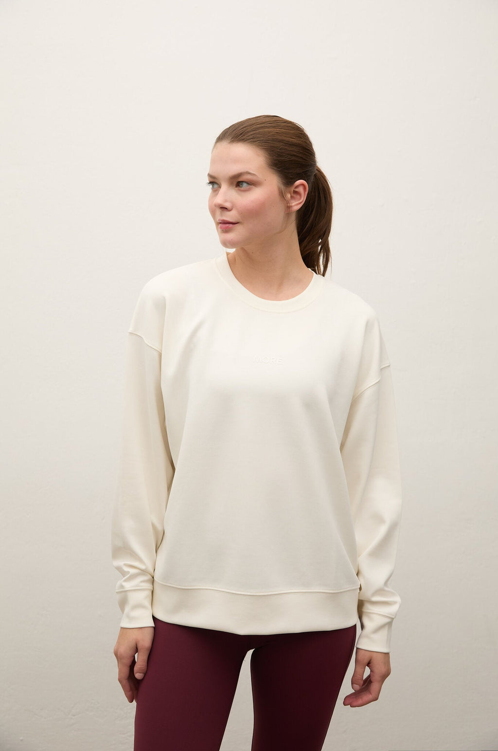 A woman wearing a cream-colored crewneck sweatshirt and burgundy leggings, standing against a plain off-white wall.