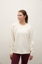 A woman wearing a cream-colored crewneck sweatshirt and burgundy leggings, standing against a plain off-white wall.
