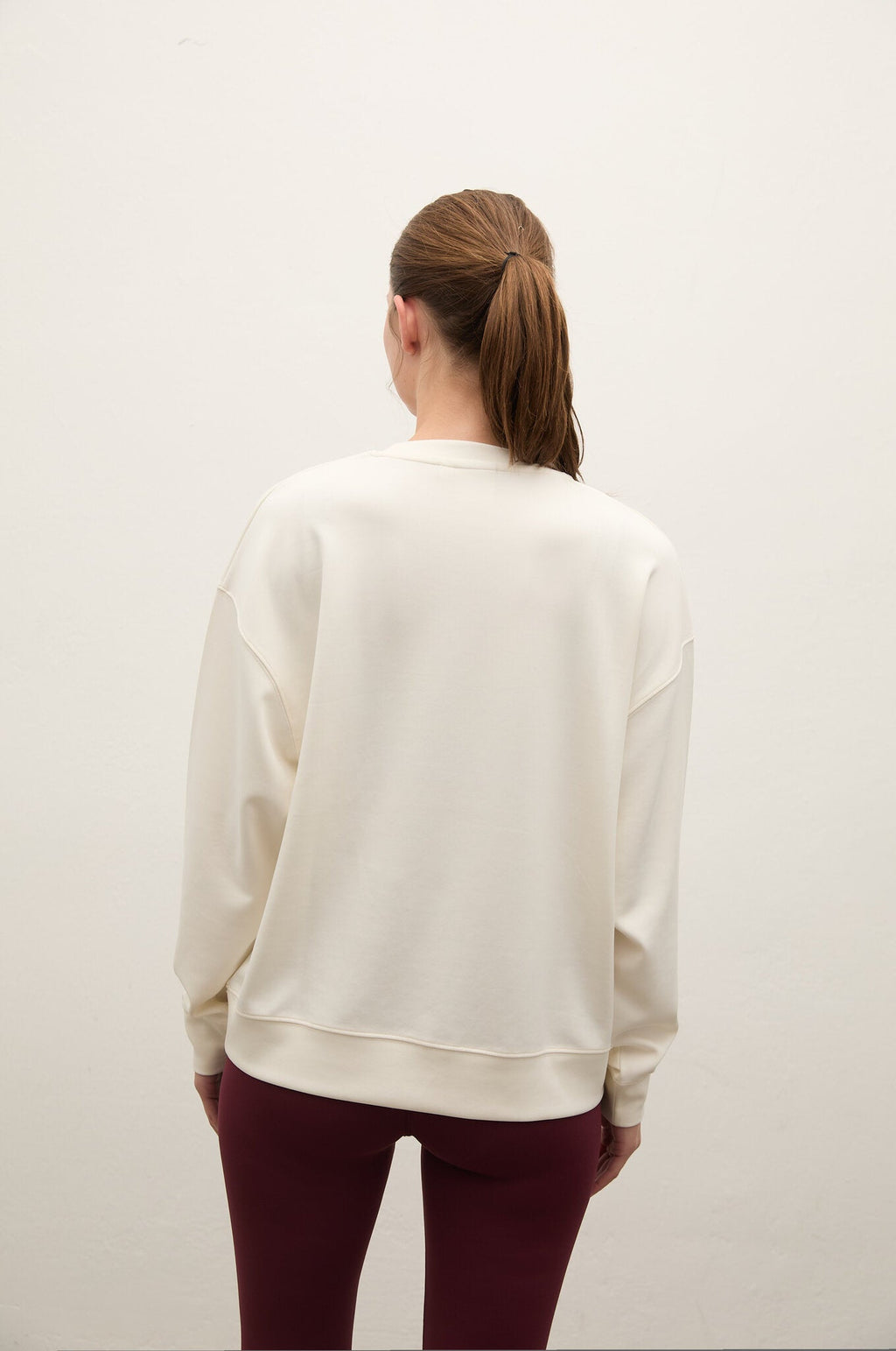 A woman viewed from behind wearing a cream-colored crewneck sweatshirt and burgundy leggings, standing against a plain off-white wall.