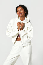 A smiling woman wearing a matching off-white cropped zip-up hoodie and jogger sweatpants, posing against a light gray studio background.