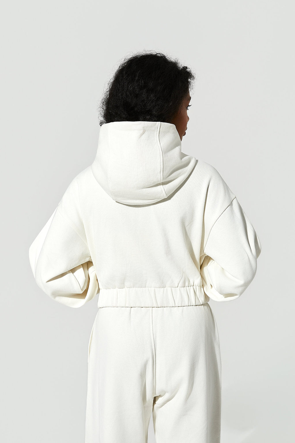 A model shown from the back wearing a cream-colored hooded sweatshirt and matching sweatpants against a light gray studio background.