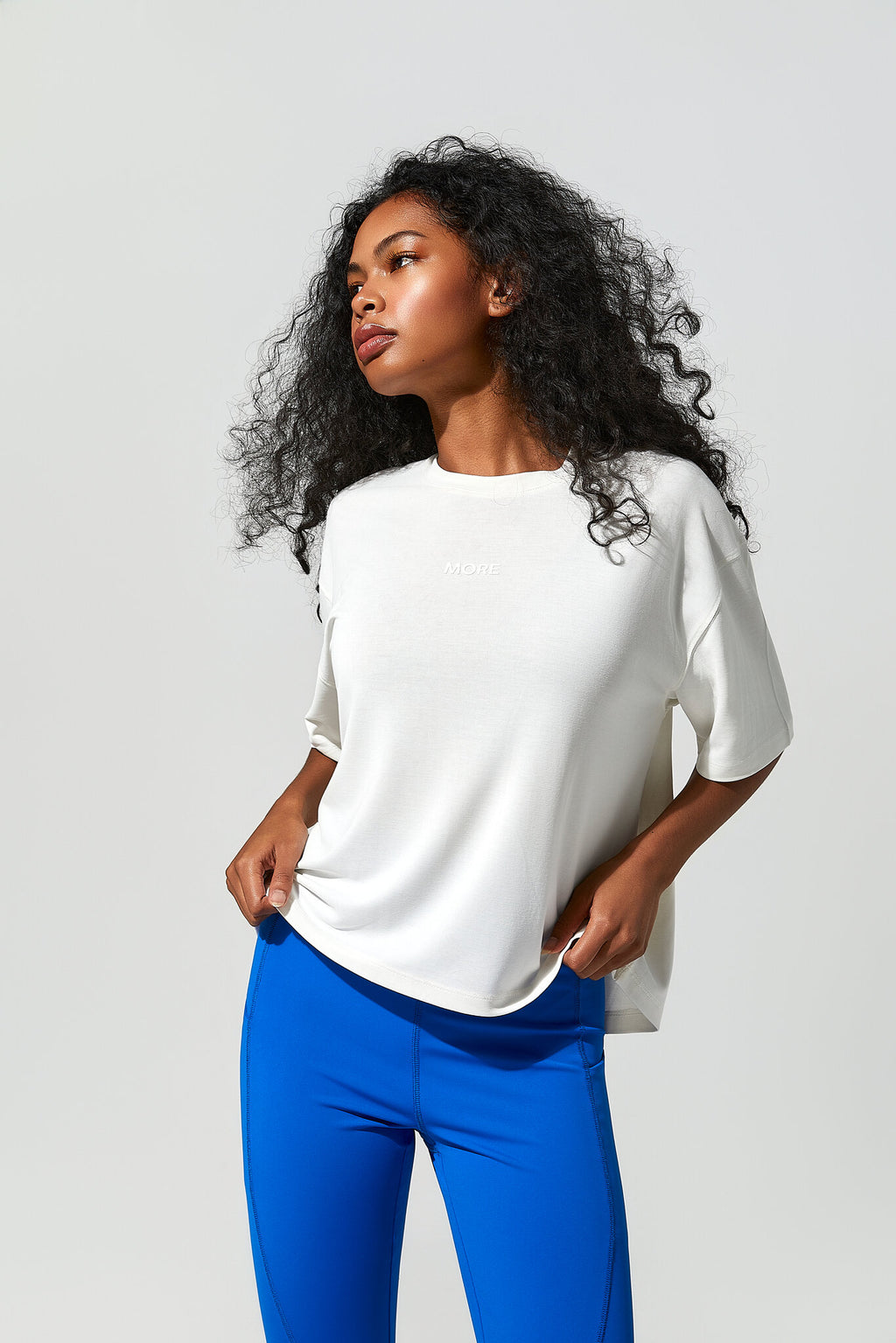 A woman with curly black hair wearing a white short-sleeve knit top and bright blue leggings, posing against a light gray studio background.