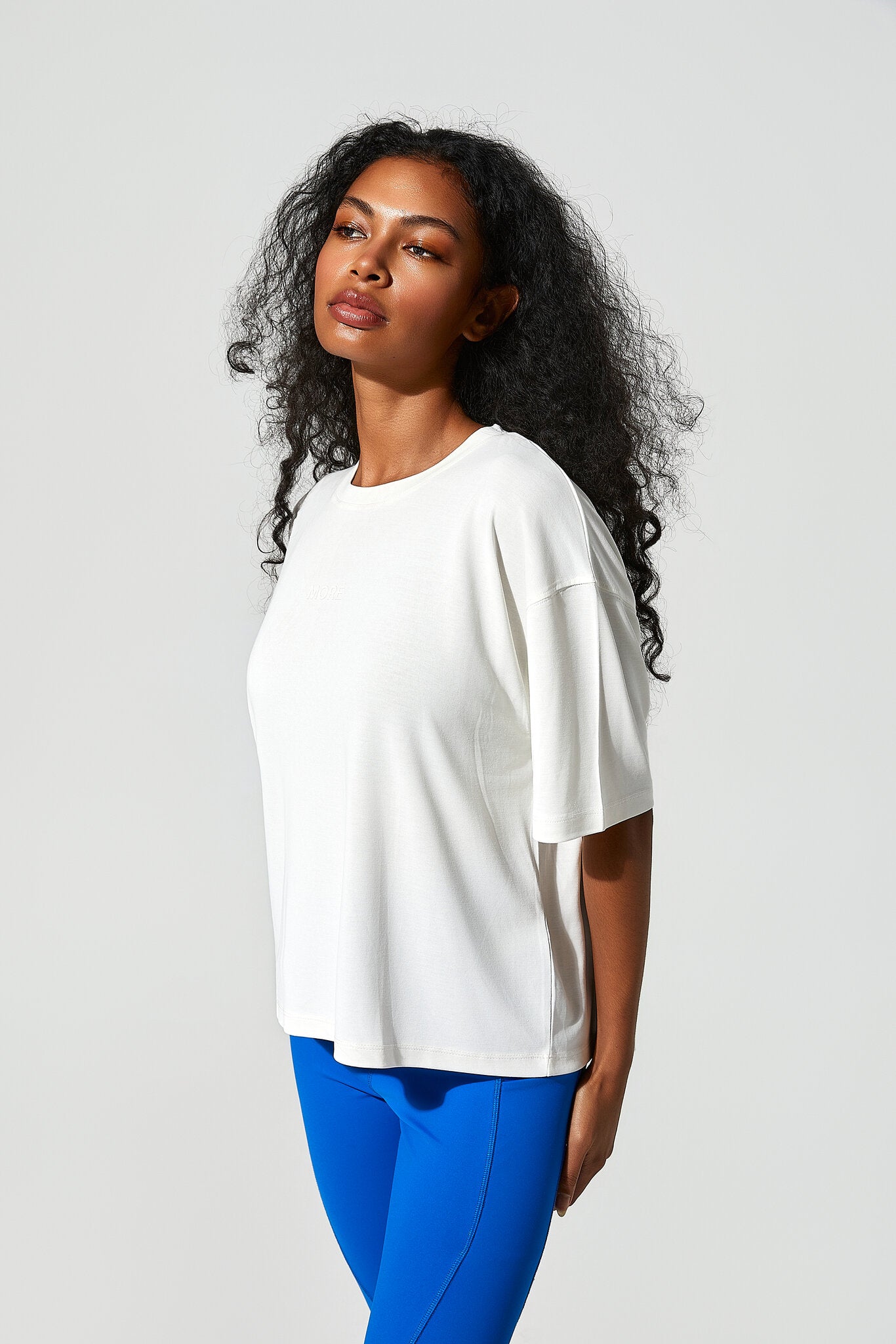 A woman with long, curly black hair is shown from the side, wearing an oversized white short-sleeve crewneck t-shirt and bright blue leggings, set against a plain light gray studio background.