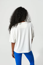 A woman with long, curly black hair is shown from behind wearing a white short-sleeve t-shirt and bright blue leggings against a light gray studio background.