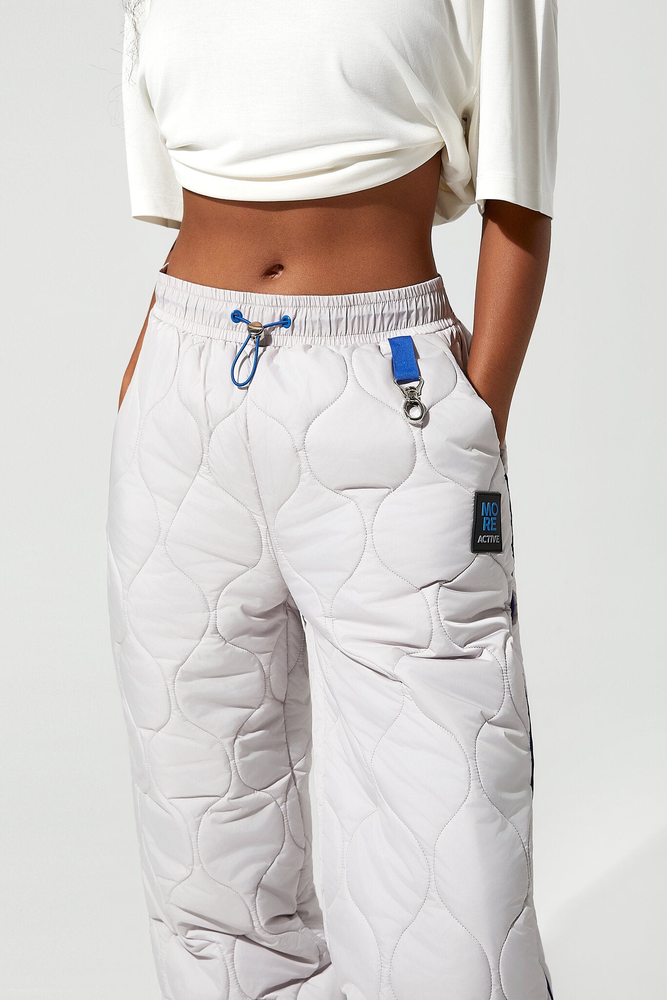 A model wearing white quilted sweatpants with a blue drawstring and a small black brand patch, paired with a white cropped top against a light gray background.