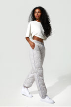 A woman modeling light gray quilted puffer pants with a thin blue side stripe, paired with a white cropped t-shirt and white sneakers, against a plain light gray studio background.