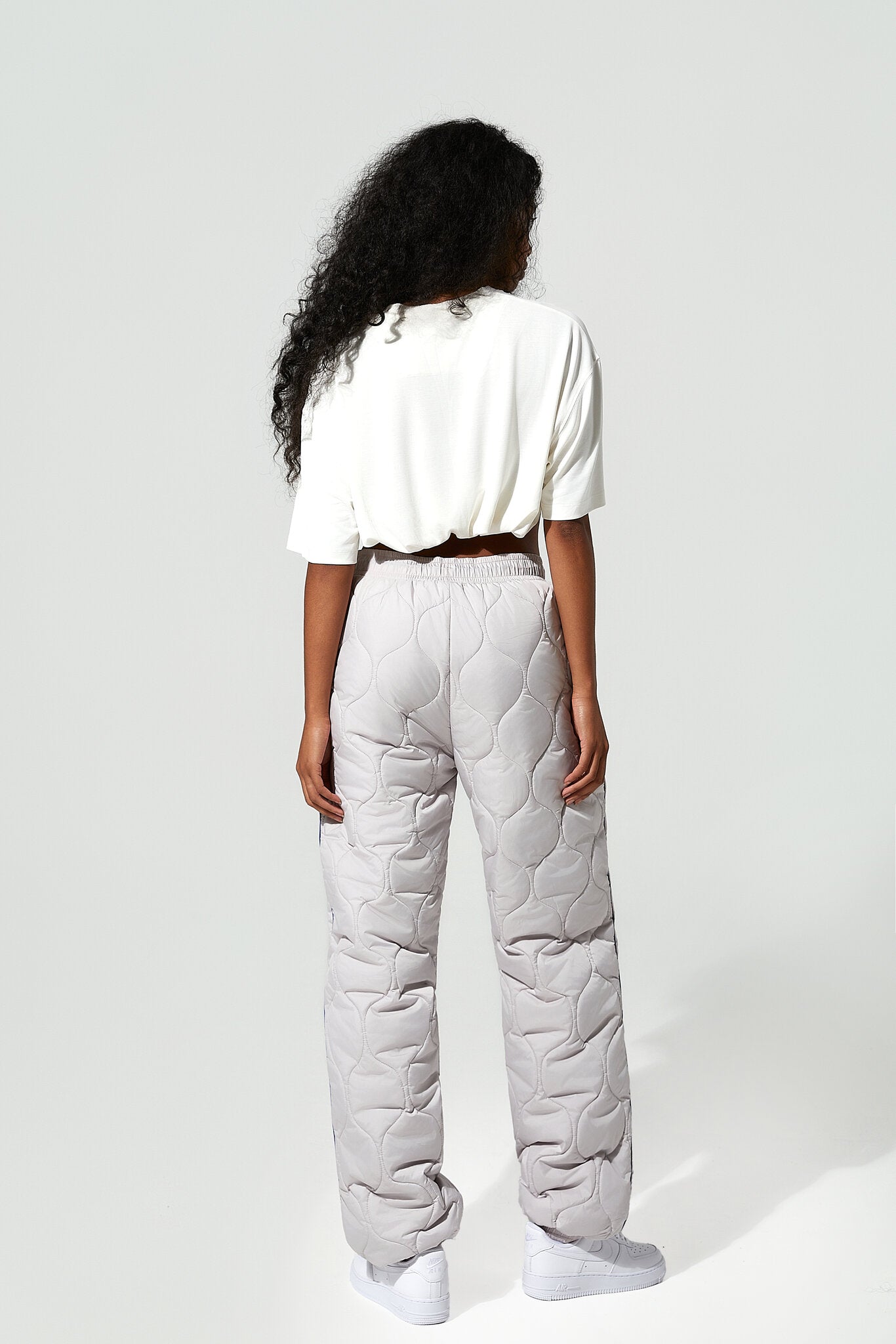 A model shown from the back wearing light gray quilted puffer pants, a white short-sleeve top, and white sneakers against a plain light gray studio background.