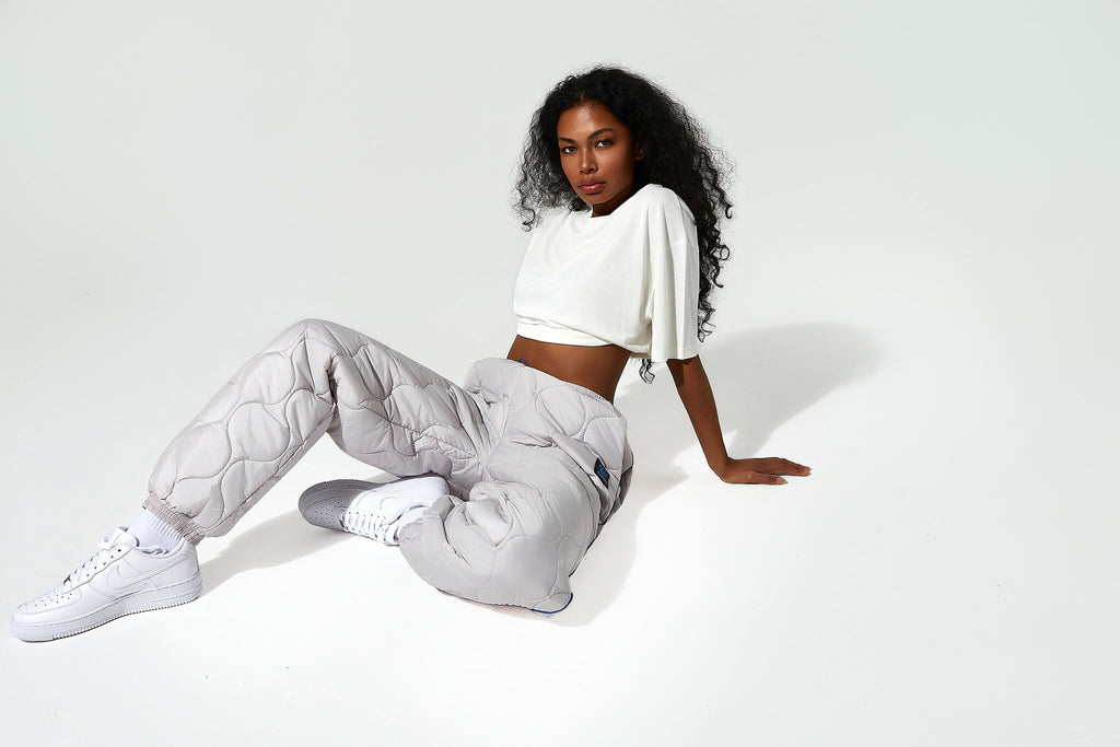 A model wearing light gray quilted sweatpants, a white cropped t-shirt, and white sneakers sits on a white studio floor against a white background.