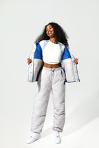 A woman modeling a matching quilted tracksuit set consisting of a white, blue, and gray color-blocked puffer jacket and matching puffer pants, styled with a white crop top and white sneakers against a light gray studio background.