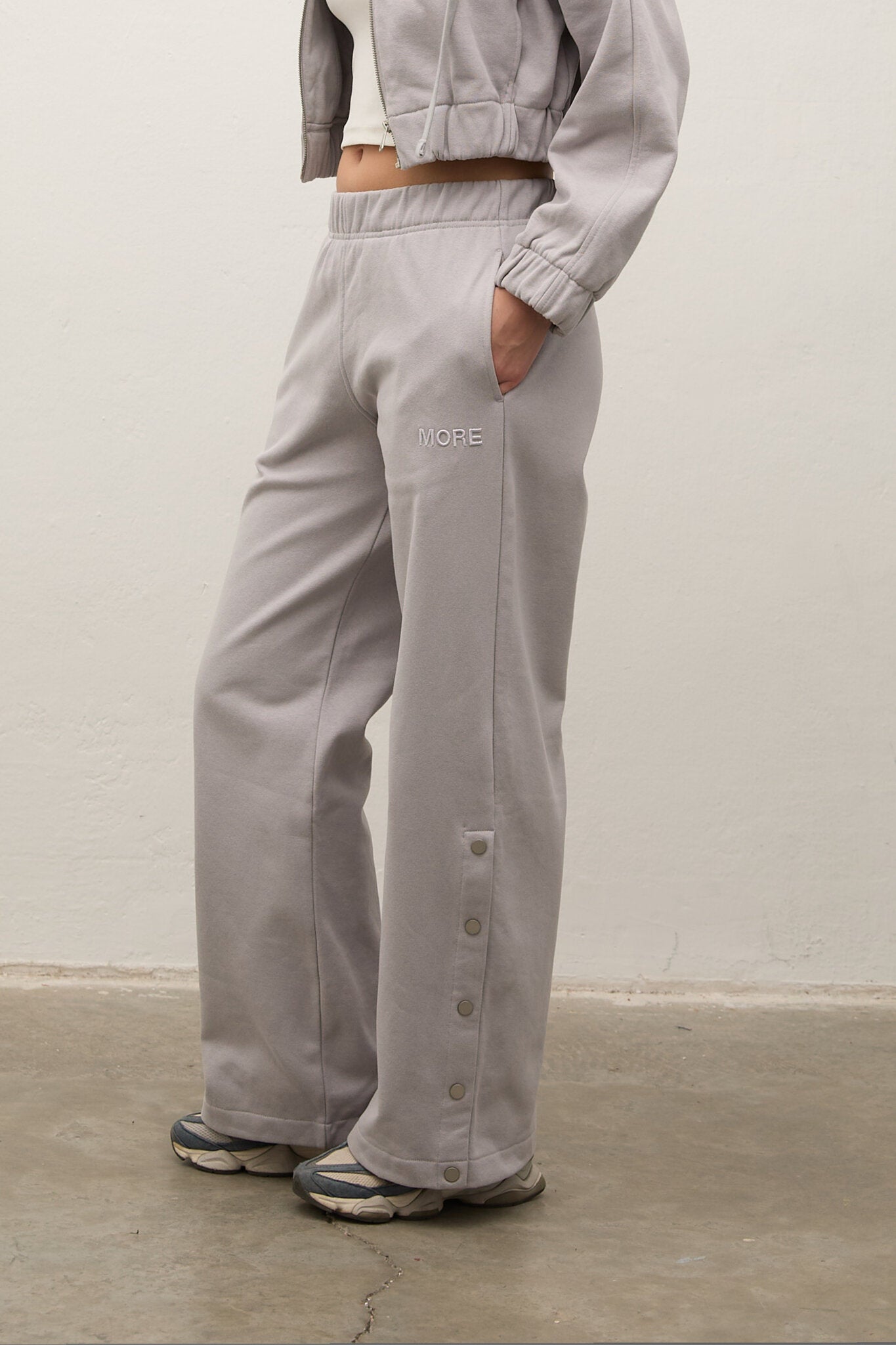 A person wearing light gray wide-leg sweatpants with side buttons and a small 'NOBE' logo on the upper thigh, paired with a matching cropped jacket and sneakers, standing against a plain off-white wall.