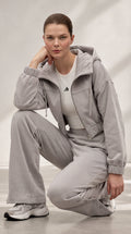 A woman crouches in a studio wearing a light gray hooded sweatshirt and matching wide-leg sweatpants over a white fitted top, with gray sneakers. A small black logo is visible on the chest.