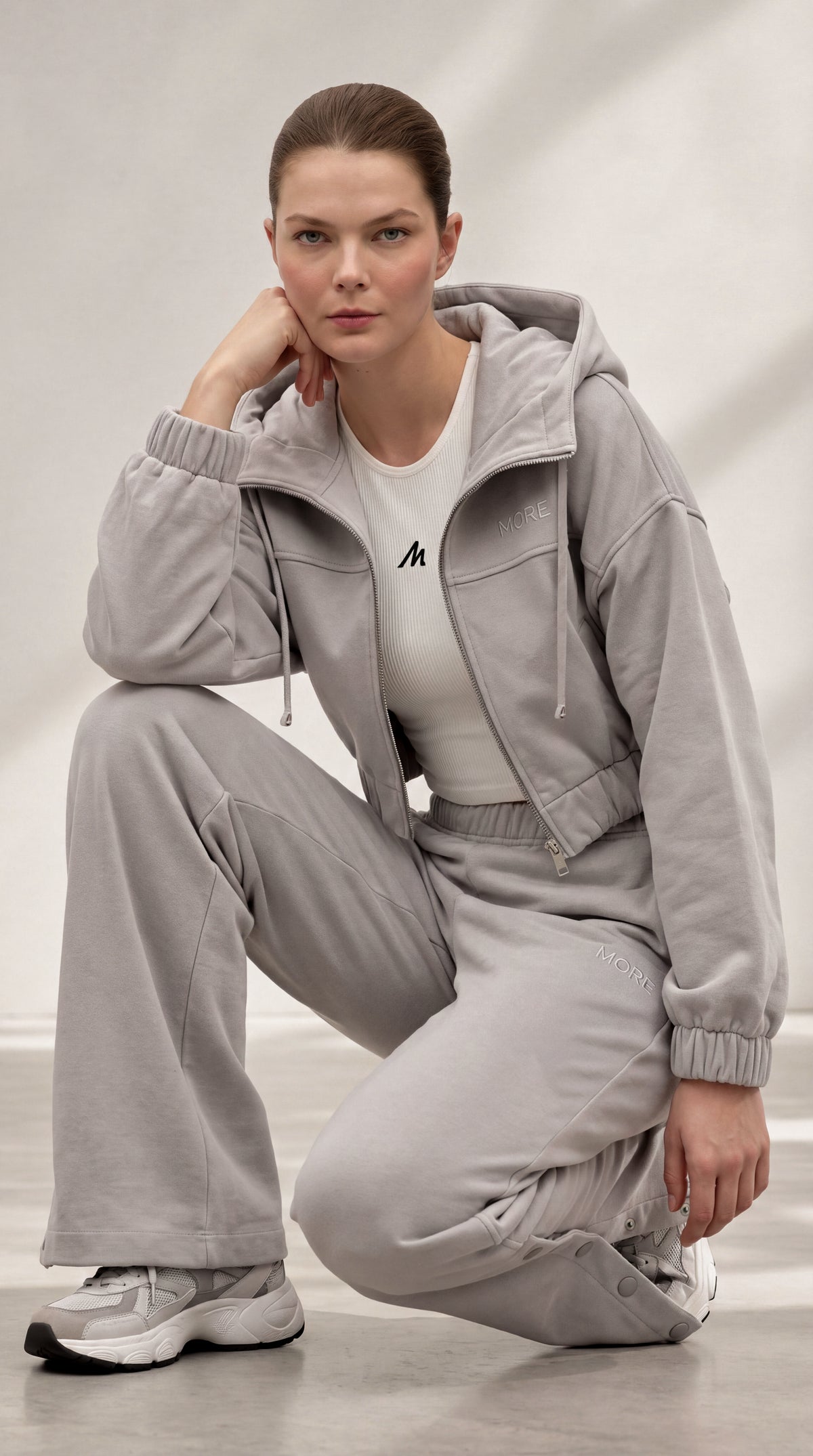 A woman crouches in a studio wearing a light gray hooded sweatshirt and matching wide-leg sweatpants over a white fitted top, with gray sneakers. A small black logo is visible on the chest.