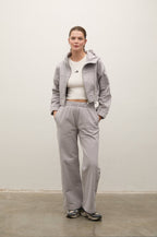 A model stands against a plain off-white wall wearing a light gray matching tracksuit consisting of a cropped zip-up hoodie and wide-leg sweatpants, styled with a white crop top and sneakers.