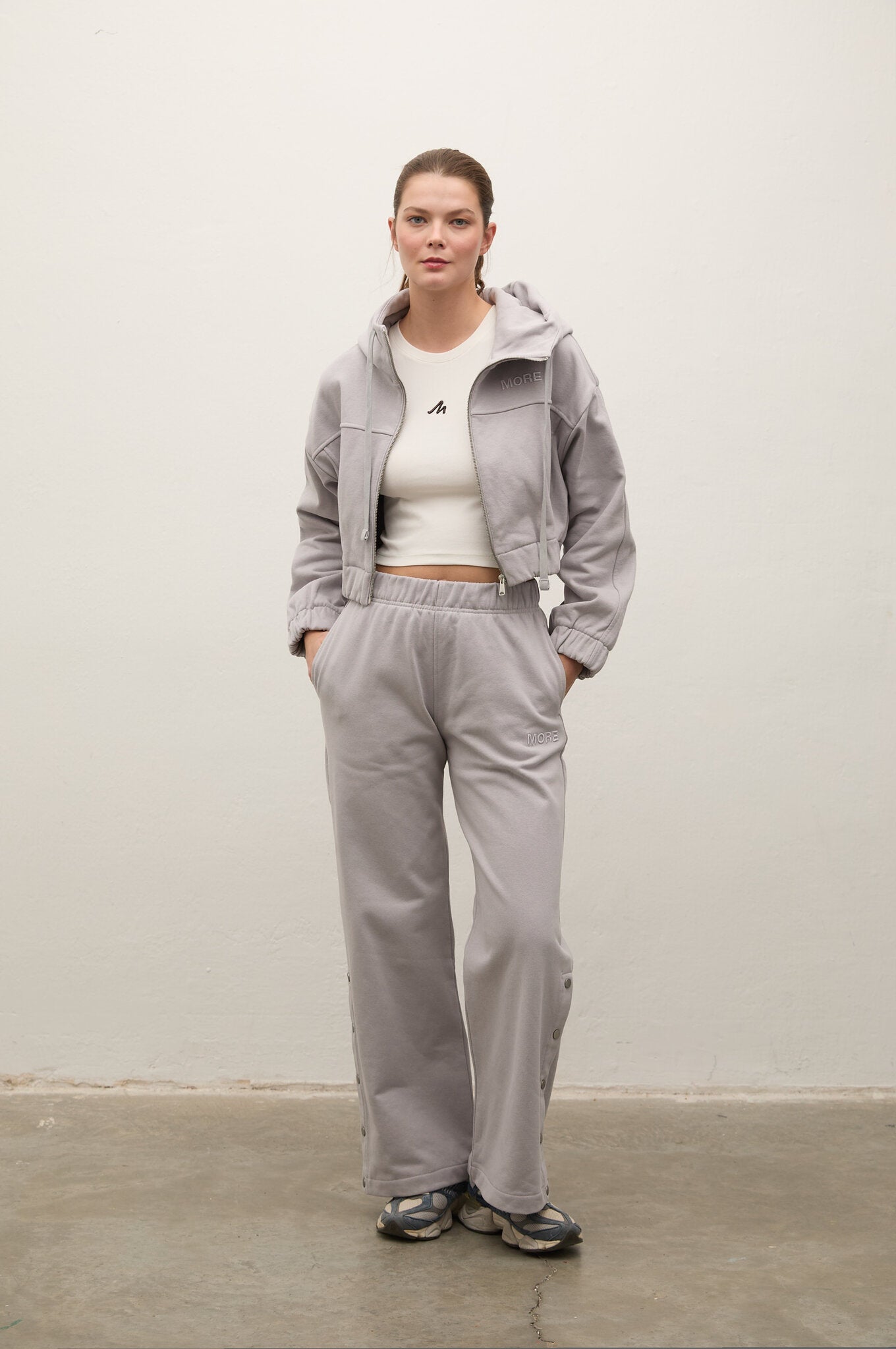 A model stands against a plain off-white wall wearing a light gray matching tracksuit consisting of a cropped zip-up hoodie and wide-leg sweatpants, styled with a white crop top and sneakers.