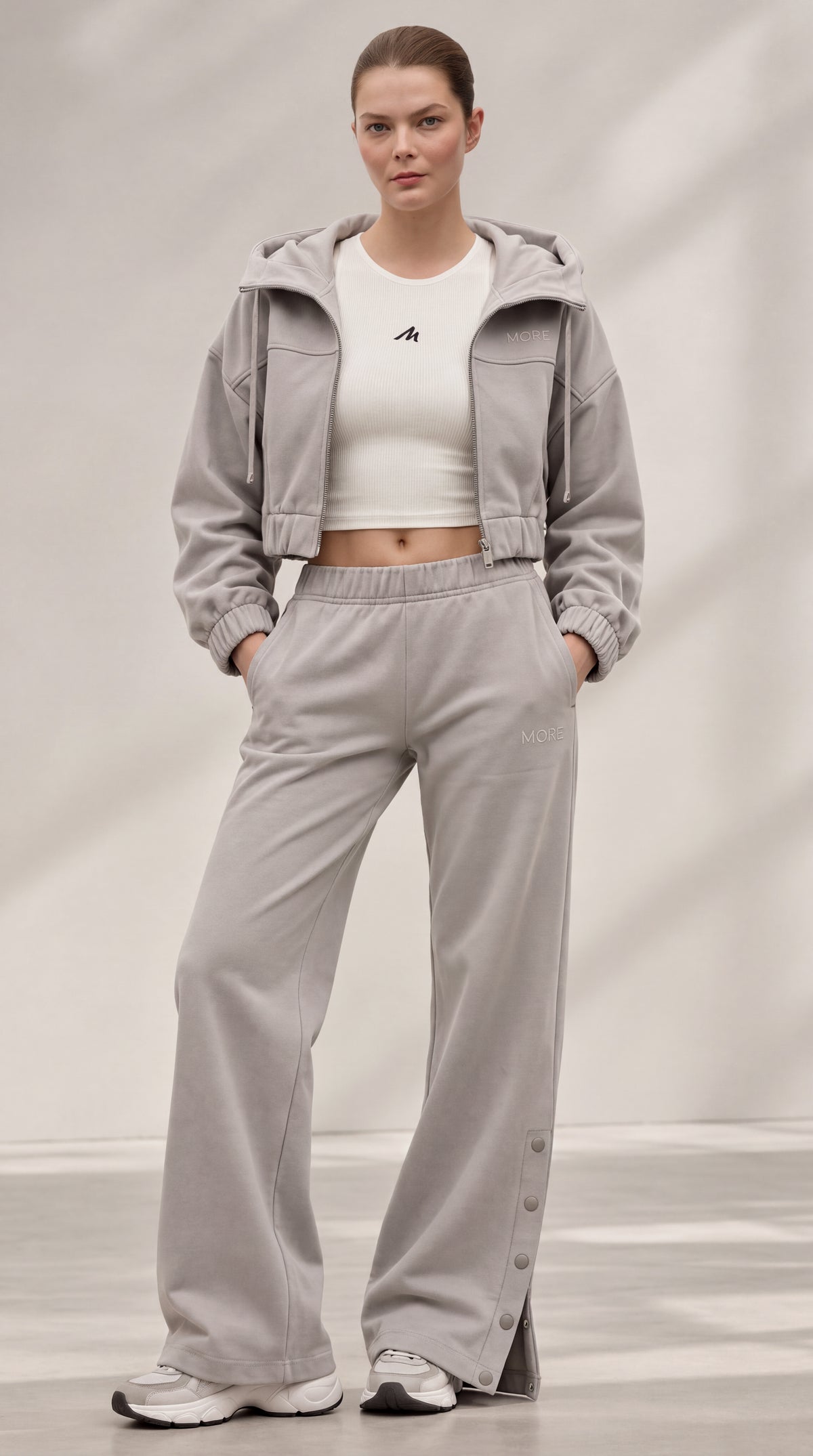 A model wearing a light gray two-piece tracksuit consisting of a cropped hooded jacket and matching wide-leg sweatpants, styled with a white cropped tank top and white sneakers.