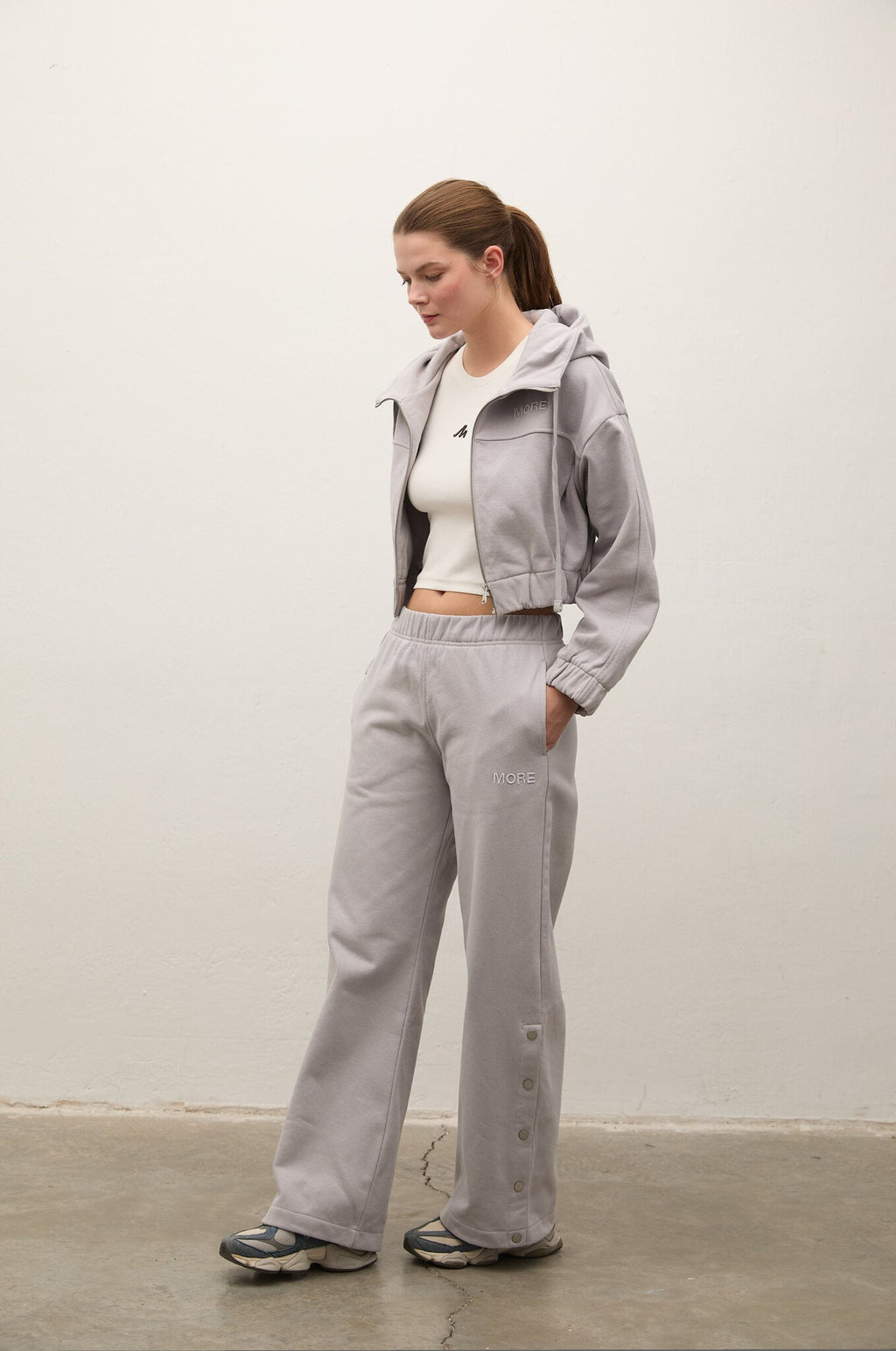 A woman modeling a light gray hooded zip-up jacket and matching wide-leg sweatpants, styled with a white cropped tank top and sneakers, standing against a plain off-white wall.