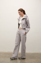 A woman modeling a light gray hooded zip-up jacket and matching wide-leg sweatpants, styled with a white cropped tank top and sneakers, standing against a plain off-white wall.