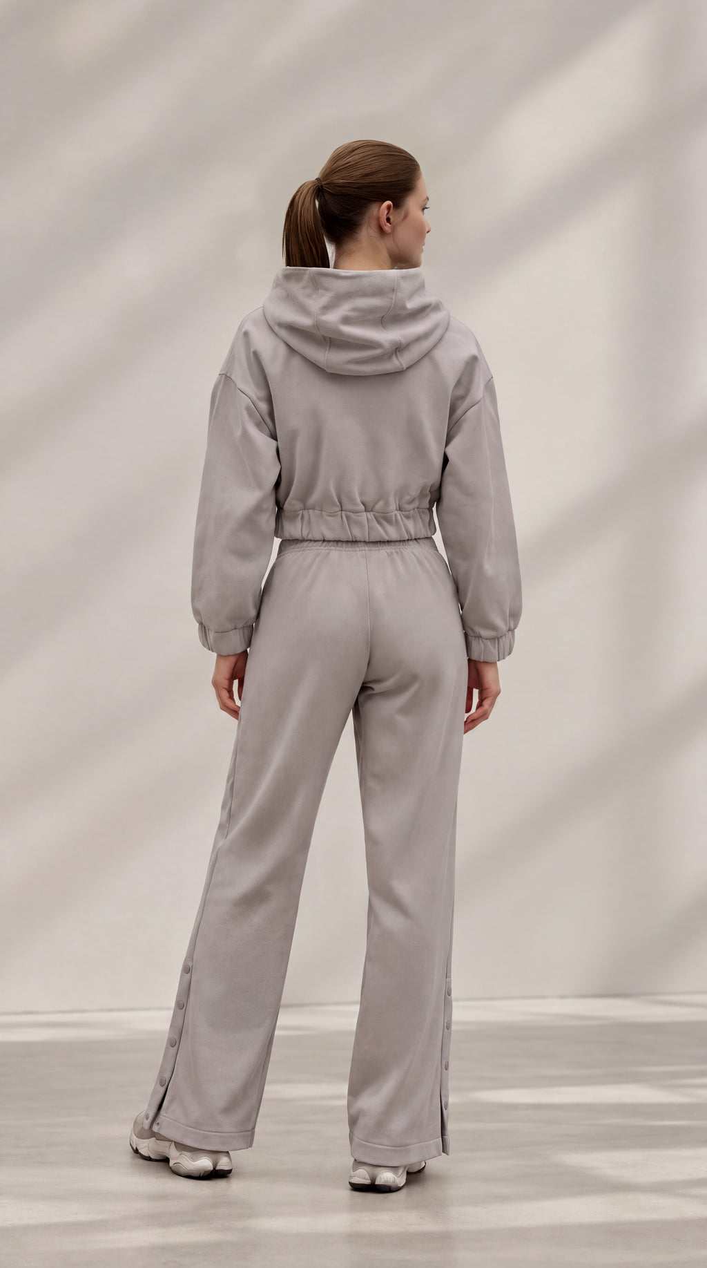 A woman viewed from behind wearing a light gray hooded jumpsuit with wide legs and side slits, standing in a bright studio with soft shadow patterns on the wall.