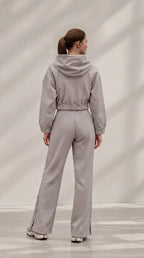 A woman viewed from behind wearing a light gray hooded jumpsuit with wide legs and side slits, standing in a bright studio with soft shadow patterns on the wall.