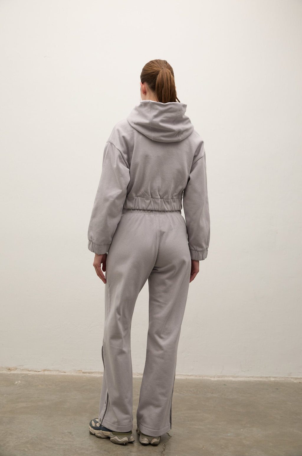 A model viewed from behind wearing a light gray hooded sweatshirt and matching wide-leg sweatpants, standing against a plain off-white wall.