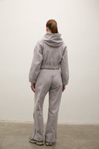 A model viewed from behind wearing a light gray hooded sweatshirt and matching wide-leg sweatpants, standing against a plain off-white wall.