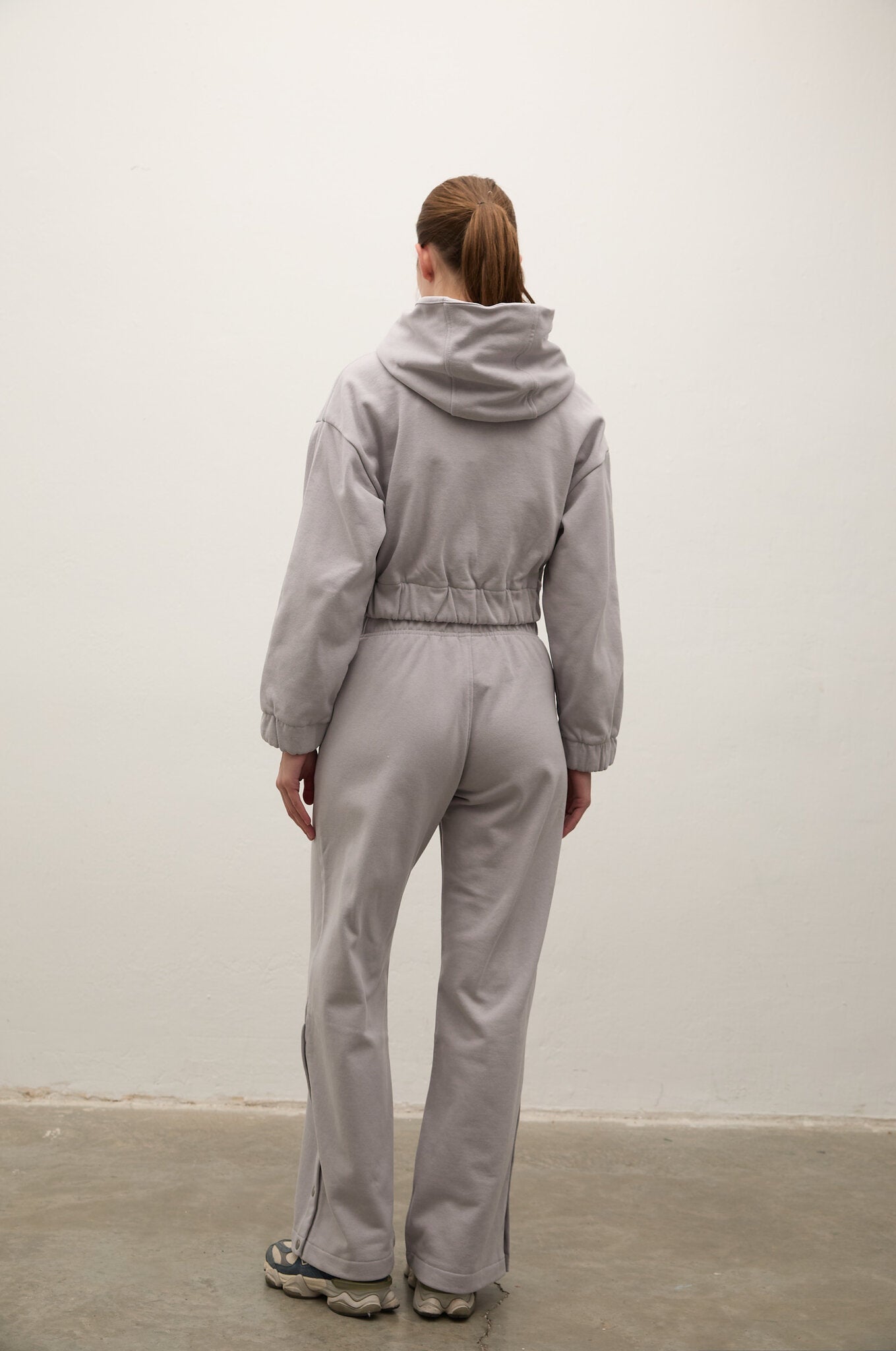 A model viewed from behind wearing a light gray hooded sweatshirt and matching wide-leg sweatpants, standing against a plain off-white wall.