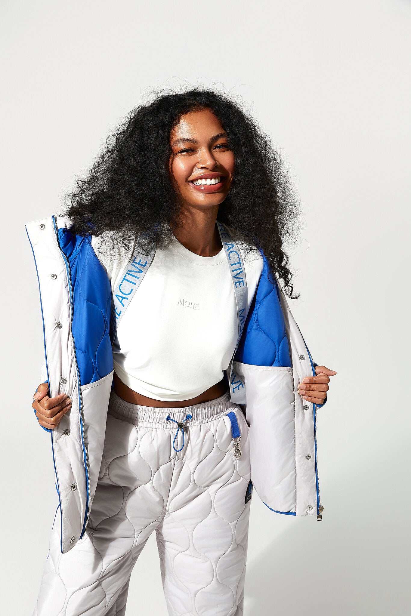 A smiling woman with curly black hair models a white, blue, and gray quilted puffer jacket and matching quilted sweatpants against a plain light gray studio background.