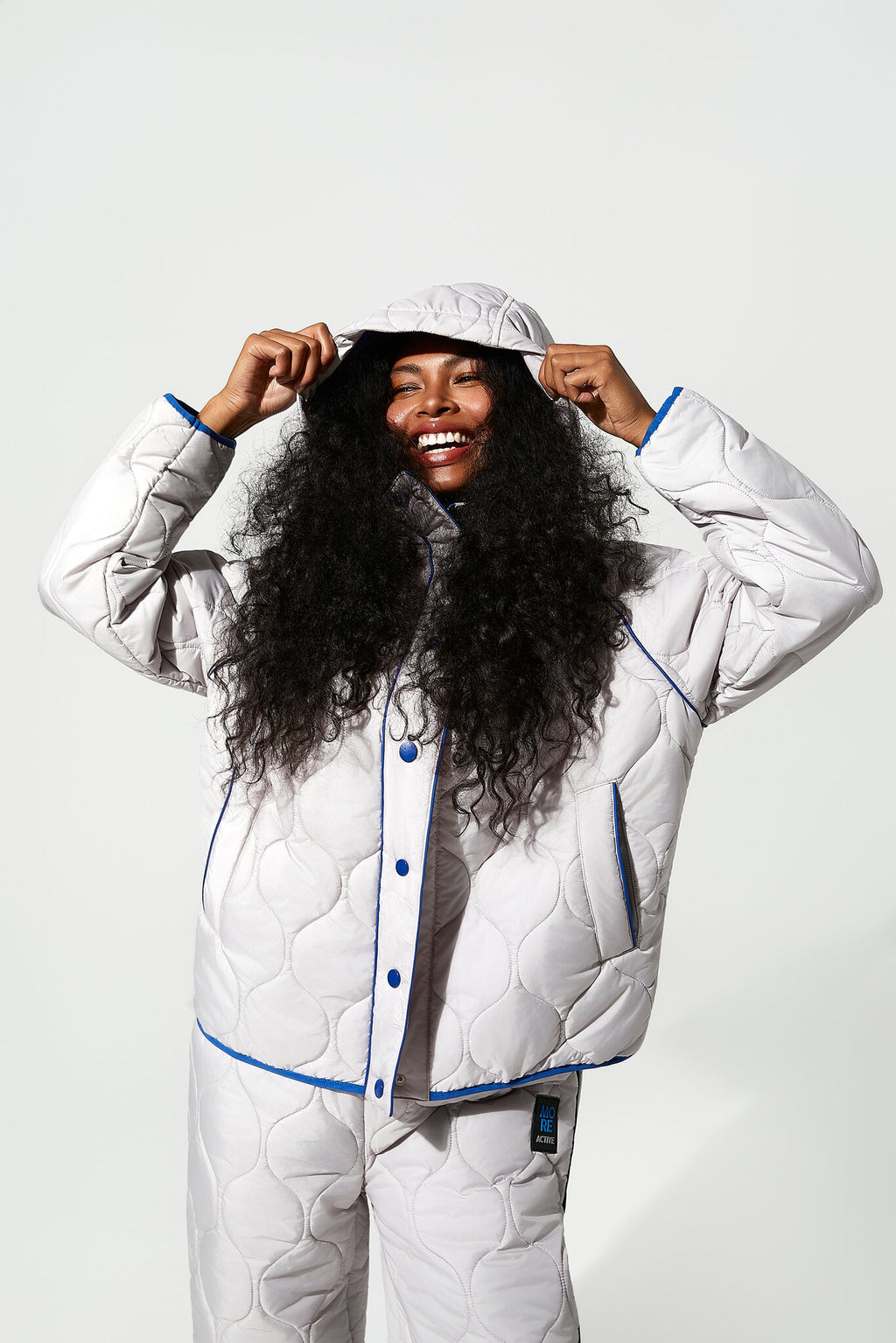 A smiling woman with long curly black hair models a matching white quilted pajama set with blue piping and snap-button front, including a hooded jacket and pants, against a light gray studio background.