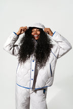 A smiling woman with long curly black hair models a matching white quilted pajama set with blue piping and snap-button front, including a hooded jacket and pants, against a light gray studio background.