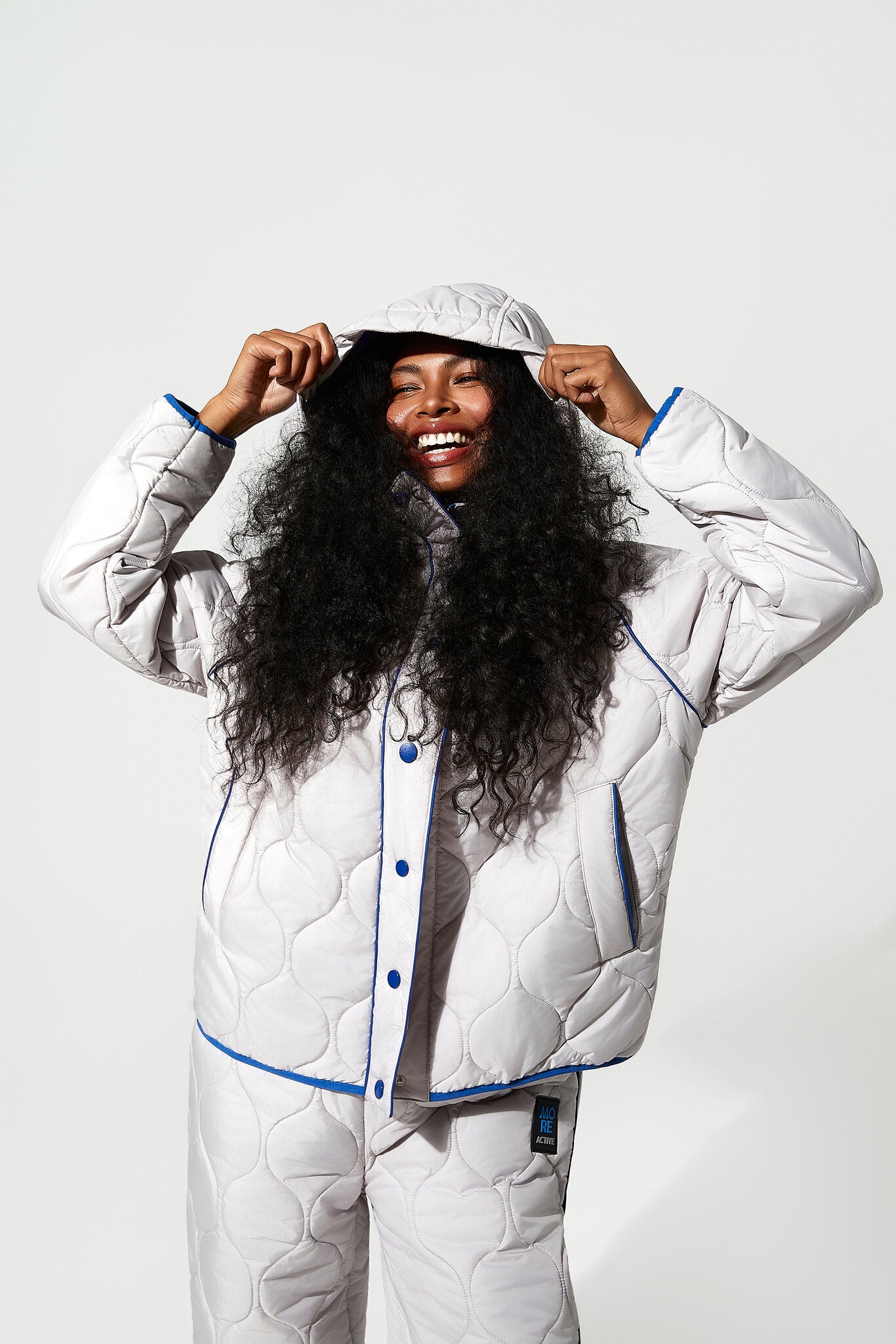 A smiling woman with long curly black hair models a matching white quilted pajama set with blue piping and snap-button front, including a hooded jacket and pants, against a light gray studio background.
