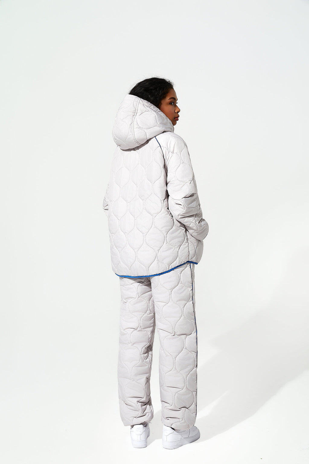 A model shown from the back wearing a light gray quilted puffer jacket and matching pants with a thin blue piping accent, set against a plain white studio background.