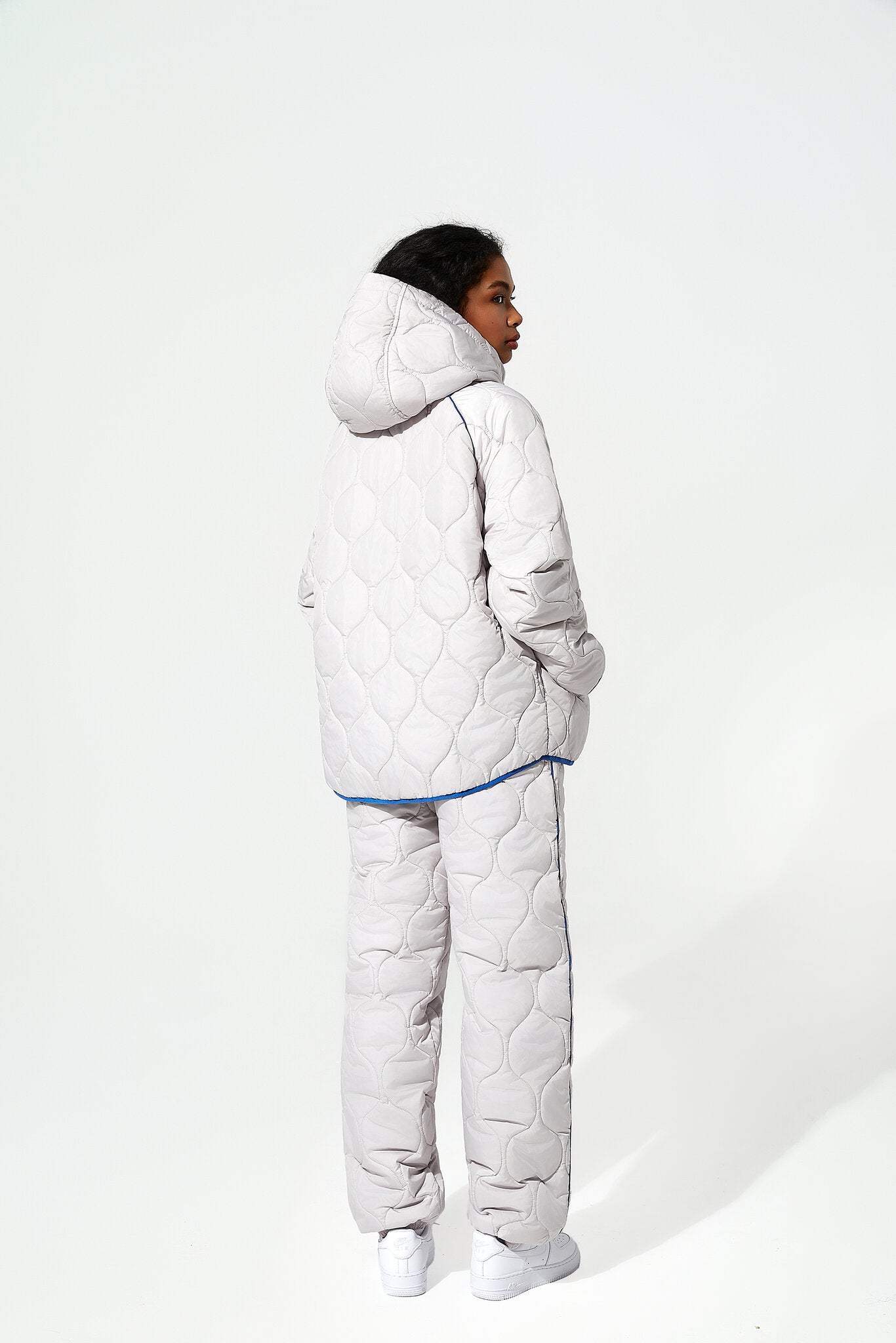A model shown from the back wearing a light gray quilted puffer jacket and matching pants with a thin blue piping accent, set against a plain white studio background.