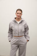 A woman modeling a light gray cropped zip-up hoodie and matching sweatpants set against a plain off-white wall.