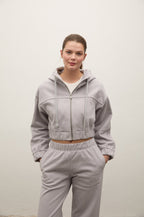 A woman modeling a light gray cropped zip-up hoodie and matching sweatpants set against a plain off-white wall.